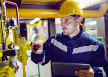 Serious dedicated plant worker in working clothes and with protective helmet on head screwing valve and holding tablet.